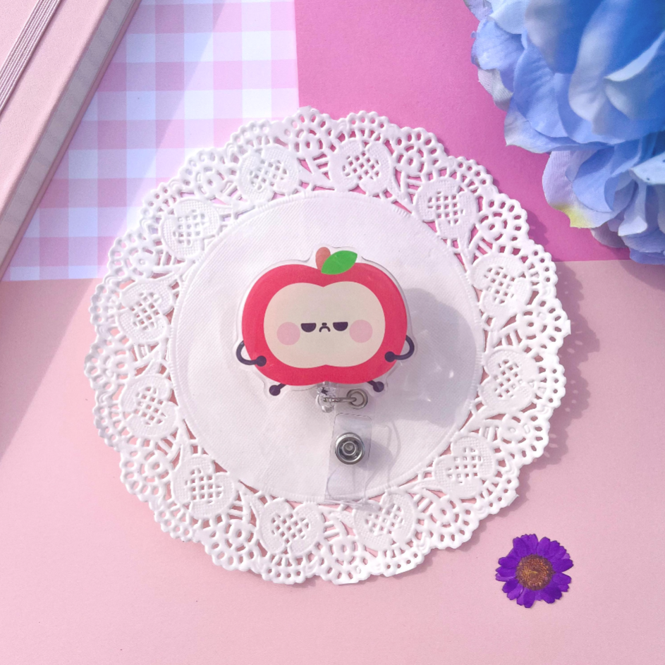 A cute red apple-shaped retractable badge reel with a cartoon face, displayed on a white lace doily over a pink background with decorative flowers.